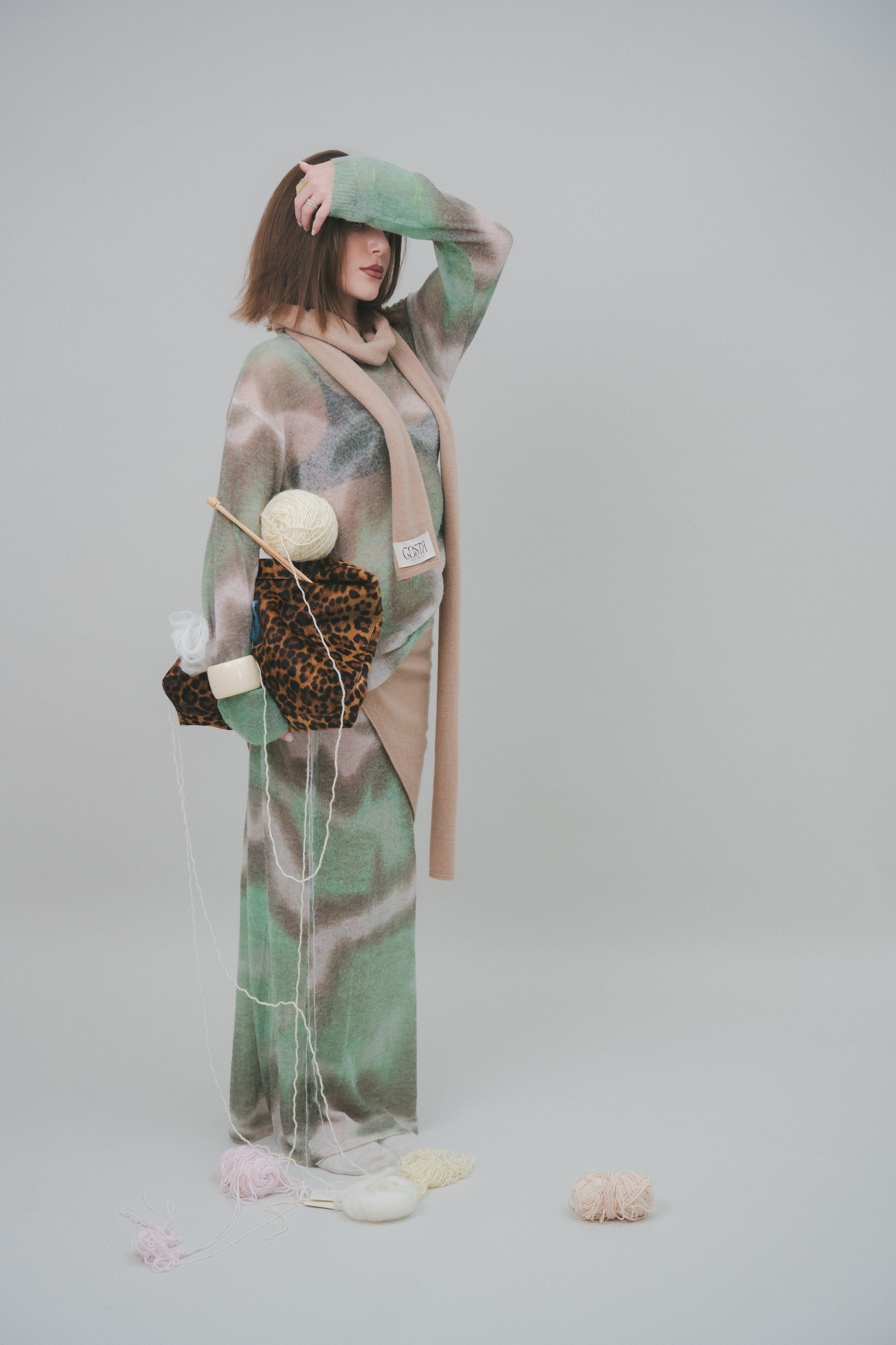 Artistic editorial shot of a model with a beige scarf and raw wool yarn balls, highlighting Costa Knit Atelier's artisanal hand-knitting heritage.