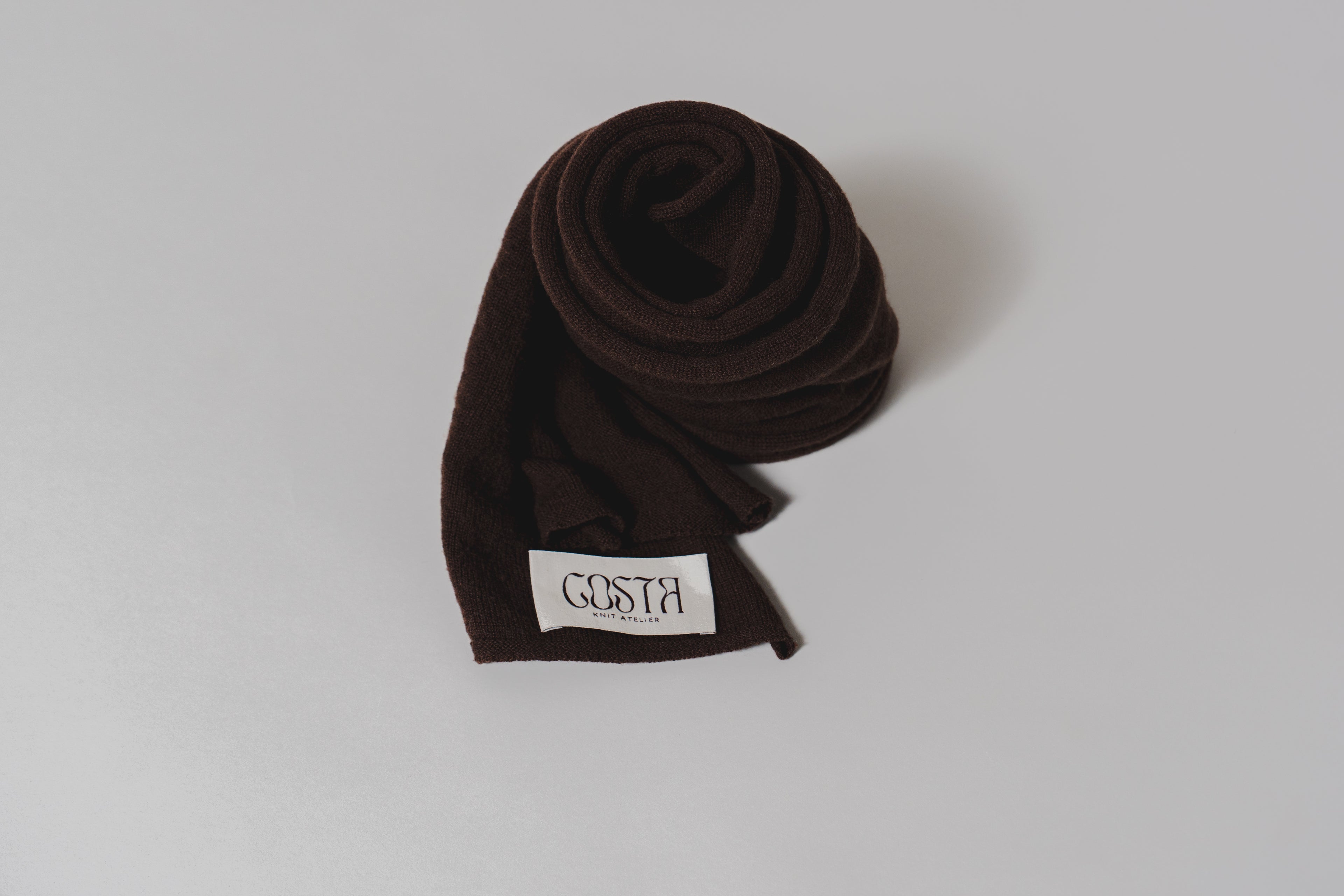 Close-up of the Costa Knit Atelier label on a luxury cashmere thin scarf in brown, highlighting our use of extra-fine natural fibers and artisanal craft.